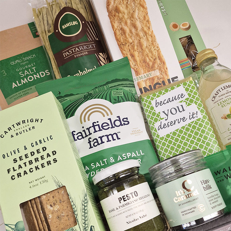Gavekurv med delikatesser som pasta, crackers, pesto og chokolade i grønne nuancer.